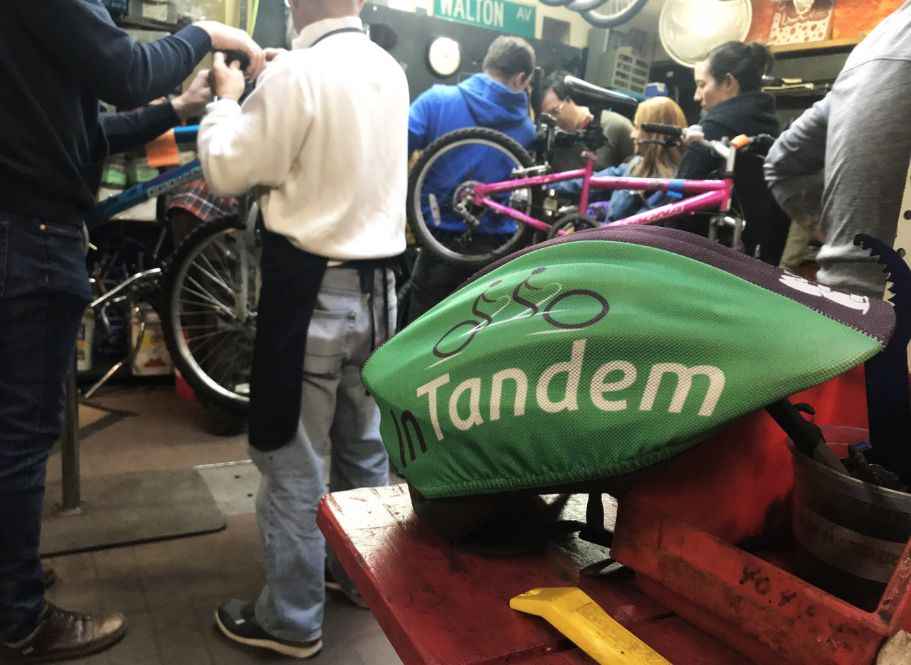 IMAGE DESCRIPTION: A bike helmet with a green InTandem helmet cover rests on a bike shop table in the foreground, and a group of people are handling bikes and tools in the background.