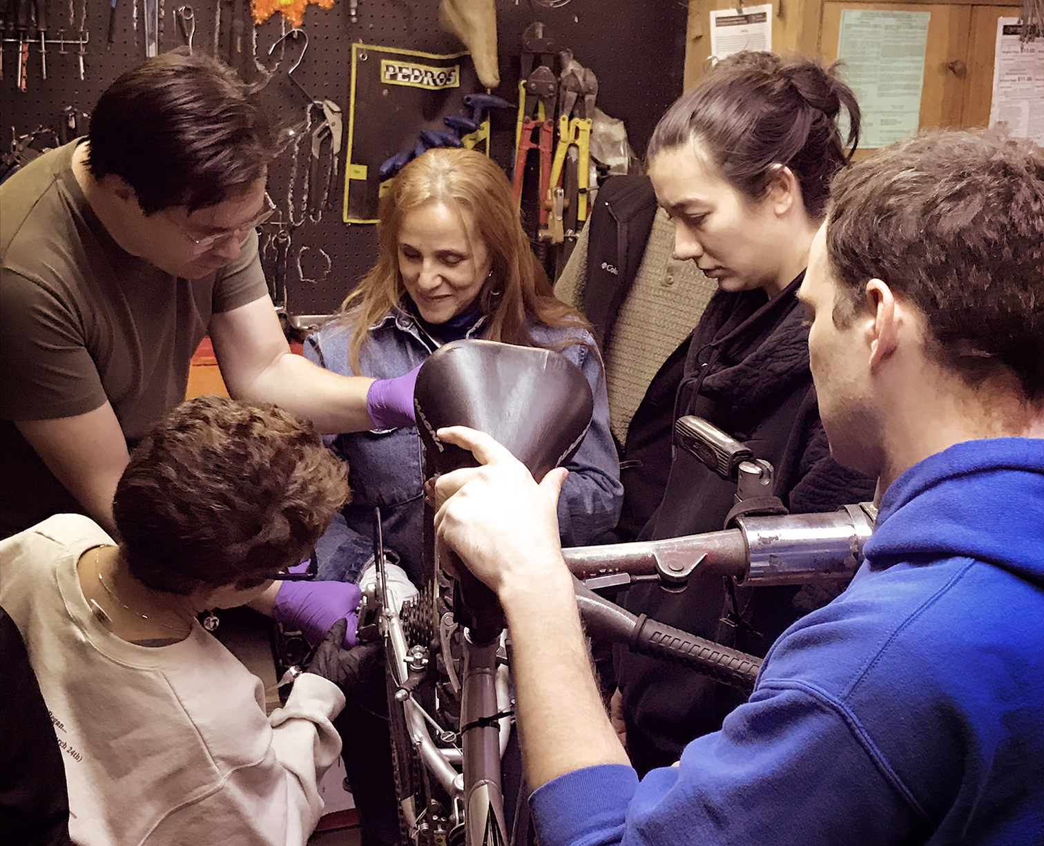 IMAGE DESCRIPTION: Two men and three women stand around the rear wheel of a bicycle, inspecting the gear system. Two are wearing nitrile gloves. The group is surrounded by a variety of tools hung on the wall.